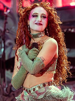 A photo of Chappell Roan live in concert. She has her arms crossed against her chest and is looking a point beyond the camera. She has white and pink full face makeup, red lipstick, and long and curly orange hair. She is wearing a green beaded cropped top and a bottom with green peacock feathers on it. She is smiling presumably at the crowd she is performing for.