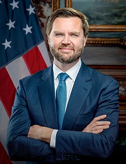 Official portrait of JD Vance, a middle-aged white man with dark hair and beard and light eyes, wearing a suit and tie, slightly grinning and crossing his arms while standing in front of an American flag.