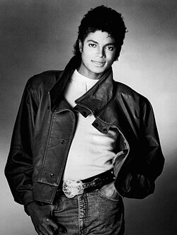 A monochrome photo of Michael Jackson posing. He wears a white shirt, jacket, gloves, and jeans with a guitar-shaped belt buckle.
