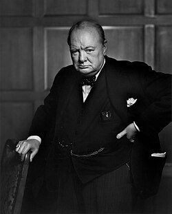 Three-quarter length portrait photograph of Churchill, 67, wearing a suit, standing and holding onto the back of a chair