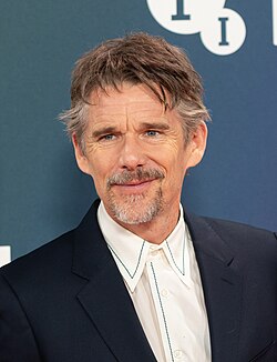 A Caucasian male with brownish-grayish hair, wearing a dark blazer and a white shirt