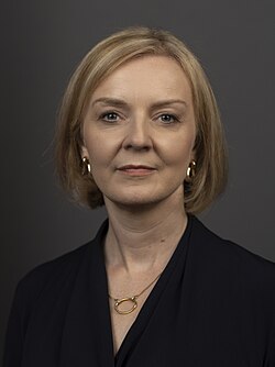 Truss facing frontward, with short blonde hair and dark clothes in front of a grey background.