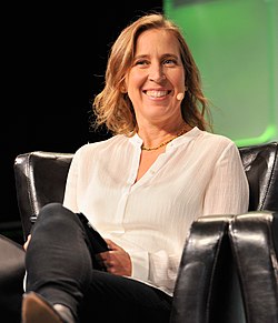 Wojcicki speaking at a TechCrunch panel. She is a middle-aged woman with blonde hair, wearing a white formal shirt and a golden necklace.