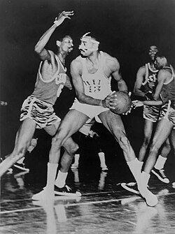 A basketball player, wearing a light-colored jersey with the number "13", is holding a basketball in front of another basketball player who is wearing a dark-colored jersey with the number "6".