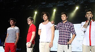 Five men stand next to each other on a stage. From left to right: Louis Tomlinson, Niall Horan, Harry Styles, Liam Payne, and Zayn Malik. Tomlinson wears bright red trousers and a white t-shirt with horizontal black stripes, and smiles while gazing at the audience. Horan, Styles, Payne, and Malik all wear identical cream trousers. Horan wears a red polo shirt. Styles wears a white t-shirt with a red rim around its neckline. Payne wears a checkered dress shirt. Malik wears a white dress shirt, with red lining around the buttons and collar. Payne has his right hand on Styles' shoulder, and Malik is the only member visibly singing.
